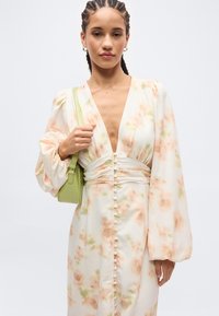 Woman with braided hair wearing a long floral dress with buttons and puffed sleeves, holding a small light green handbag on her shoulder.