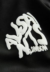 Čierna tkanina s výrazným bielym logom a textúrovaným, nepravidelným písmom, ktoré znie "ALSA NTS LONDON." Detailný pohľad.