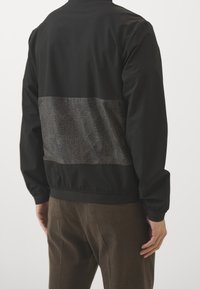 Muže oblečeného v černé bundě s texturovaným tmavě šedým panelem přes horní část zad a hnědými kordovým kalhotami, stojícího proti jednoduchému pozadí.