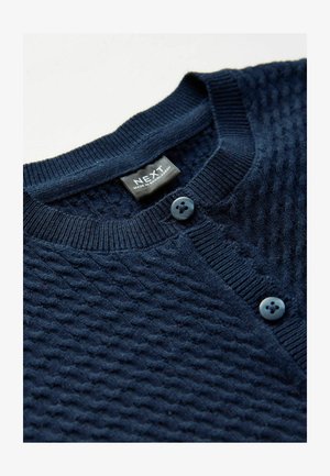 Cardigan tricotat de culoare bleu marin, cu un model texturat în diamant, având guler rotund și două butoane albastre în față. Eticheta brandului este vizibilă.