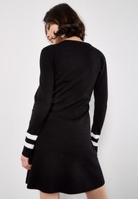 Schwarzes Kleid mit langen Ärmeln, das weiße Streifenakzente an den Bündchen aufweist. Weicher Stoff, taillierter Oberteil, der in einen ausgestellten Rock übergeht.