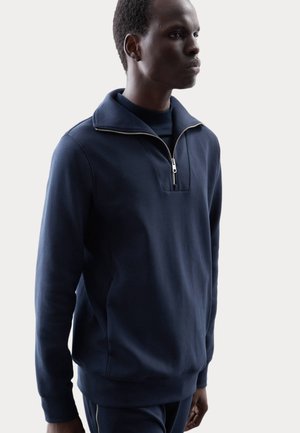Felpa blu navy con colletto alto, mezza zip e taglio boxy. Realizzata in tessuto morbido, presenta polsini e orlo a coste.