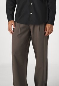 Man in een donkerbruin geplooide broek en een zwarte lange mouw knoopshirt, staand met de handen ontspannen langs de zij.