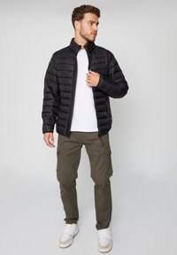 Chaqueta de plumas negra con un cuello alto, con textura acolchada y cierre de cremallera, combinada con pantalones cargo oliva y zapatillas blancas.