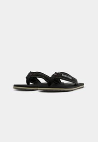 Des tongs noires avec une semelle intérieure texturée, arborant un logo sur la bride et des coutures blanches contrastantes. Semelle en caoutchouc avec une légère courbure.