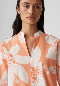 Lichte koraalkleurige blouse met een bloemenpatroon in wit en oranje. Heeft een mandarin-kraag en een gedeeltelijke sluiting bij de hals.