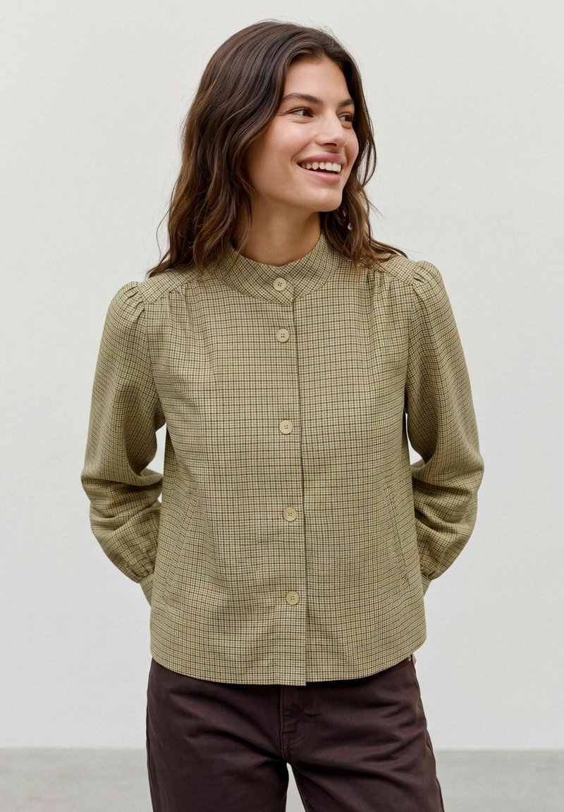 Femme souriante, portant un chemisier beige à carreaux avec boutons, épaules bouffantes et col haut, mains derrière le dos, sur fond uni.