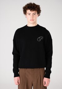 Junge Person mit lockigem Haar, die einen schwarzen Pullover mit einem kleinen gestickten Logo auf der Brust und braunen gefältelten Hosen trägt.