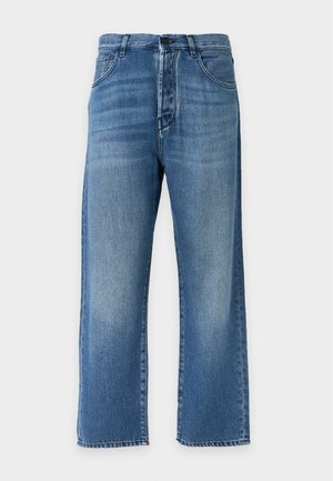 Femme portant une veste en jean avec une broderie florale rouge, un jean bleu, des bottines noires et un haut blanc, debout avec une main dans la poche.