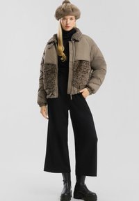 Kobieta ubrana w taupe'owy puchowy płaszcz z akcentami z sztucznego futra, pasujący futrzany kapelusz, czarne szerokie spodnie i czarne masywne buty, stoi z ręką w kieszeni.