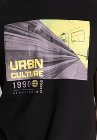 Svart t-shirt med ett grafiskt mönster i lila och gult. Texten lyder "URBN CULTURE 1990" bredvid en tågillustration.