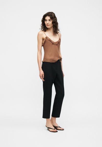 Femme aux cheveux bouclés portant un haut marron sans manches à volants, un pantalon court noir et des sandales noires à talons, debout devant un fond uni.