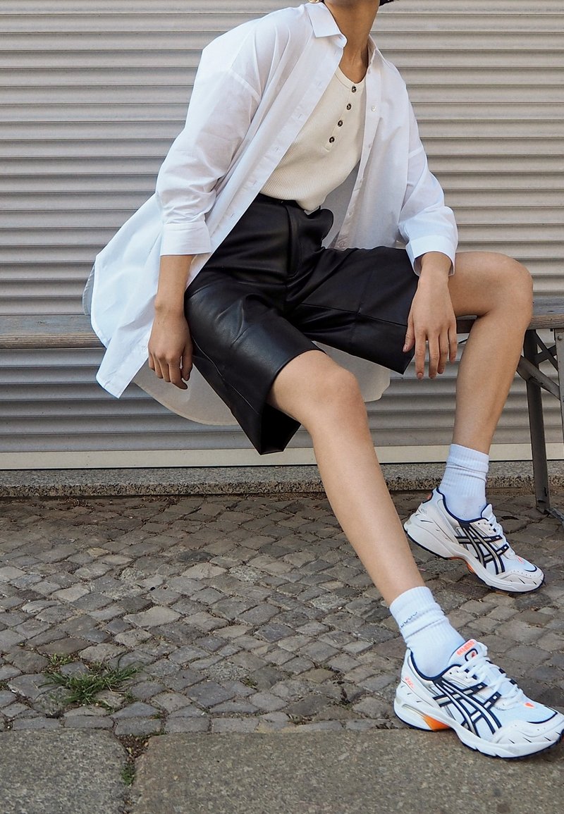 Vit överstor skjorta, beige linne, svarta lädershorts, vita och marinblå sneakers med orangea detaljer, sittande på en stenytan.