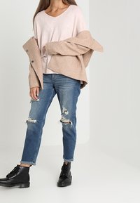 Ljusrosa v-ringad stickad topp, beige oversized jacka, slitna blå jeans och svarta snörade ankelskor med en kraftig sula.