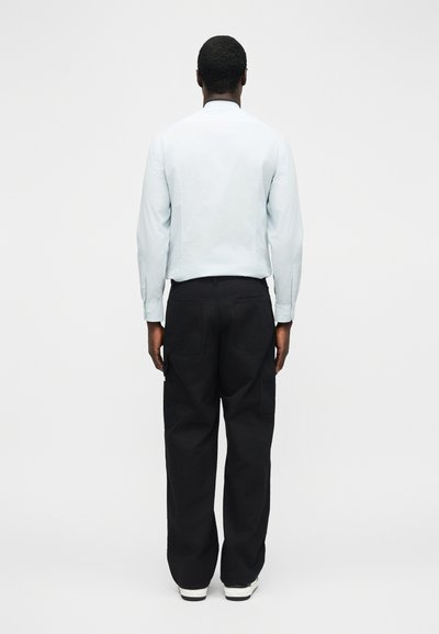 Άνδρας με το πρόσωπο στραμμένο προς τα πίσω, φορεί ένα ανοιχτό μπλε μακρυμάνικο πουκάμισο, μαύρο παντελόνι τύπου cargo με πίσω τσέπες και λευκά αθλητικά παπούτσια, σε λευκό απλό φόντο.