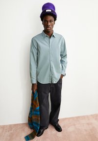 Lichtgroen langarmshirt, donkergrijze wijde broek, zwarte schoenen, houdt een multicolor gebreide trui vast, draagt een paarse gebreide muts.