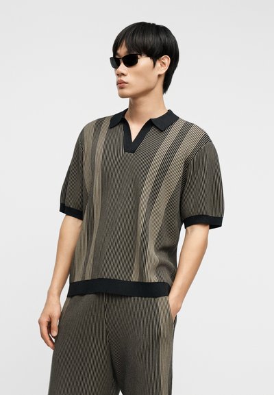 Νεαρός άνδρας με μαύρα γυαλιά ηλίου και συνδυασμό μπεζ και μαύρης κάθετης ριγέ κοντομάνικης μπλούζας και παντελονιού, με το χέρι στην τσέπη, κοιτώντας αριστερά.