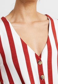 Červený a bílý pruhovaný top s výstřihem do V, s detailem knoflíků vpředu. Hladká, lehká látka.