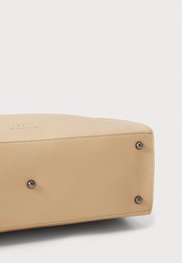 Beige leren tasbodem met afgeronde hoeken, voorzien van zes metalen noppen ter ondersteuning, en subtiele stiksels langs de randen.