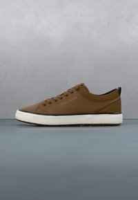 Baskets basses marron avec semelle blanche, bordure noire, lacets et logo Tommy Hilfiger sur le côté, présentées sur fond gris.