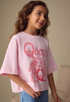 Junges Mädchen mit welligem Haar, das ein locker sitzendes pinkfarbenes T-Shirt mit rotem "Queen"-Logo und blaue Jeans trägt, steht drinnen vor einem neutralen Hintergrund.