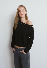 Joven con cabello largo y rubio que lleva un suéter negro de hombros descubiertos con ojales plateados y jeans grises con tachuelas a lo largo de las costuras laterales.