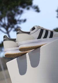 Weiße und schwarze Adidas-Sneaker mit Gummisohlen, die auf einer weißen geneigten Oberfläche im Freien mit verschwommenen Bäumen im Hintergrund liegen.