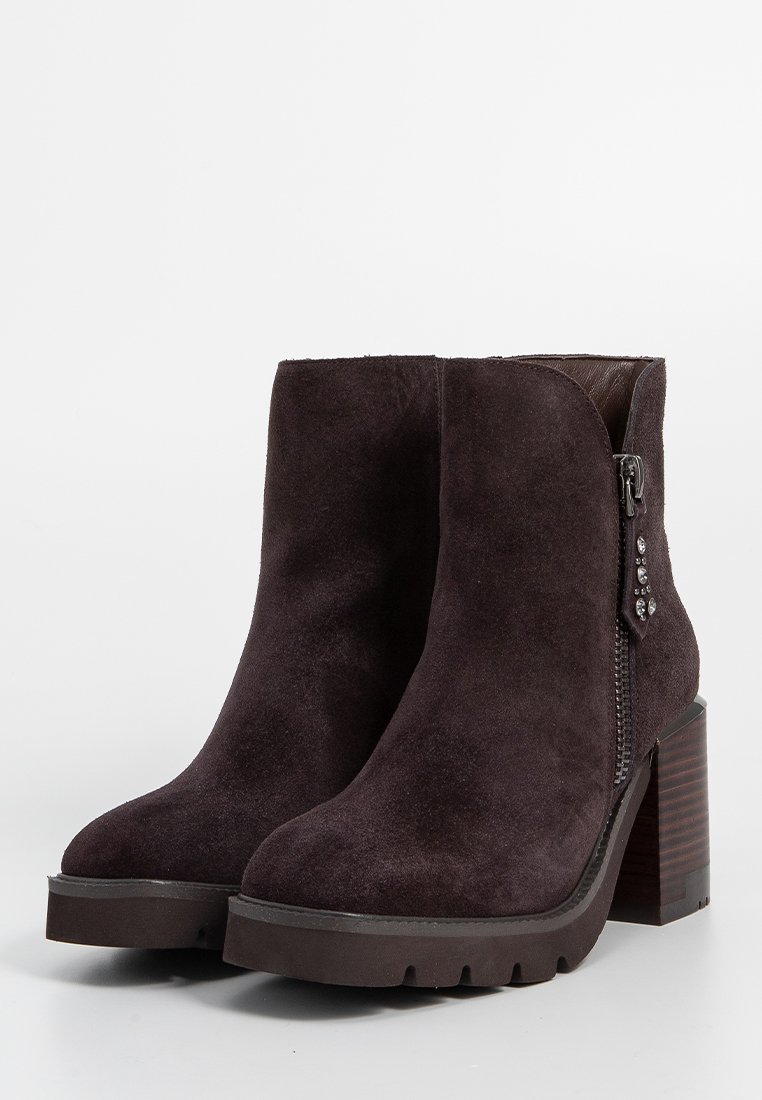 Botines de ante morado oscuro con tacón cuadrado grueso, cremallera lateral y tachuelas decorativas en el tacón. Parte superior texturizada y suela resistente.
