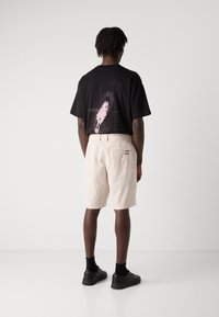 Svart t-shirt med grafisk tryck på ryggen, kombinerad med ljusbeige shorts med en patchficka och logodetalj. Svarta skor och strumpor fulländar looken.