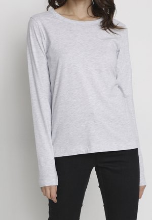 Vrouw draagt een lichtgrijs shirt met lange mouwen en zwarte broek, staand tegen een effen lichte achtergrond.