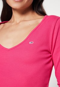 Růžový žebrovaný top s dlouhým rukávem a hlubokým výstřihem ve tvaru V, s malým značkovým logem na levé straně hrudi. Hladká textura.