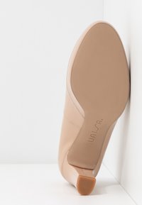 Chaussure basse en cuir beige avec talon carré, semelle tournée vers l'avant, appuyée contre un mur d'angle blanc.