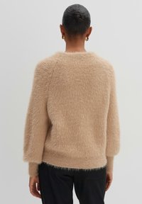 Personne avec les cheveux en chignon porte un pull beige duveteux et un pantalon noir, vue de dos sur un fond clair uni.