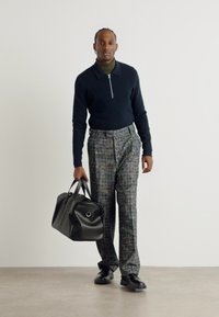 Pulover în navy închis cu fermoar, pulover verde cu guler înalt, pantaloni gri cu model, geantă de voiaj din piele neagră și pantofi negri. Texturi netede, croială ajustată.