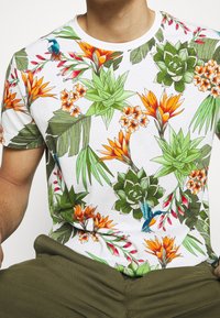Camiseta blanca de manga corta con estampados tropicales vibrantes que incluyen hojas verdes, flores naranjas y amarillas, y un detalle de colibrí.