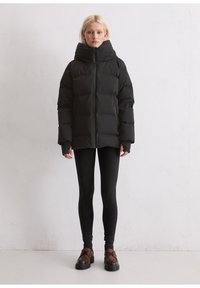 Frau, die eine schwarze Daunenjacke mit hohem Kragen, schwarze Leggings, dunkle Socken und braune Slipper trägt, steht vor einer weißen Wand.