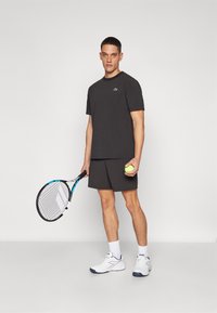 Svart treningst-skjorte og shorts, står med en tennisracket og ball. Skoene er hvite med marineblå detaljer. Enkel, sporty design.