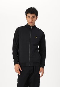 Giacca nera con zip e colletto alto, caratterizzata da pannelli testurizzati, polsini elasticizzati e un piccolo logo giallo sul petto.