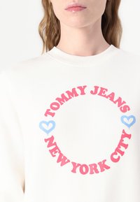Hvit hettegenser med rund grafikk som har "TOMMY JEANS" i fete røde bokstaver og "NEW YORK CITY" under, aksentuert av blå hjerter.