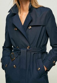 Marinblå trenchcoat med dubbelknäppning, knappar i guldton, bältesmidja och sidofickor. Tillverkad av slätt, vattenavvisande tyg.