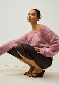 Różowy sweter z dzianiny w wzór diamentowy i cekinami, zestawiony z brąską spódnicą. Modelka stoi w pozycji przysiad na jasnym tle, nosząc czarne sandały na koturnie.