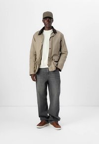 Beige jas met donkere kraag, witte trui, donkergrijze spijkerbroek, bruine sneakers en een geruit petje. Casual pasvorm met gestructureerd ontwerp en zakken.