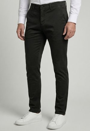 Homme portant un pantalon noir slim-fit, une chemise blanche à manches longues et des baskets blanches, debout devant un fond uni.