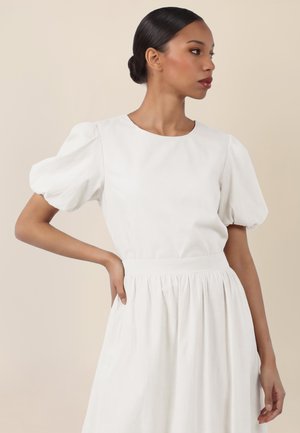 Frau mit dunklem Haar zu einem Dutt frisiert, trägt ein weißes Kleid mit Puffärmeln, schaut zur Seite und hat eine Hand in die Hüfte gestützt.