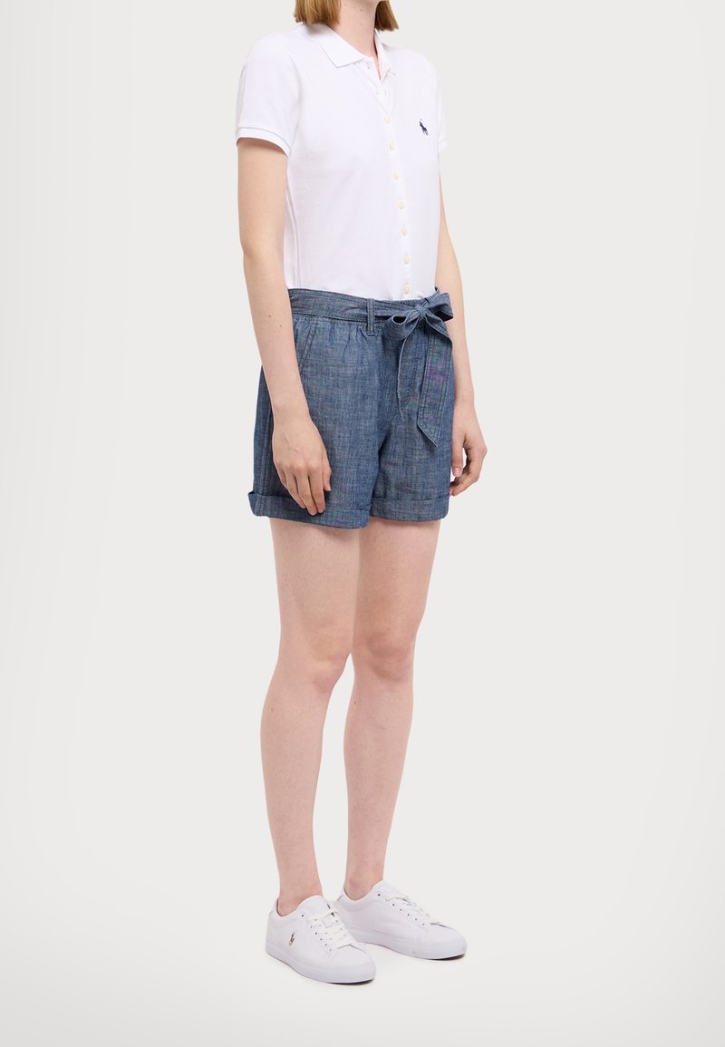 Kvinna iklädd vit kortärmad knappad polo, blå högmidjade shorts med knytning och vita sneakers, stående mot en neutral bakgrund.