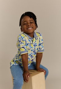 Poloshirt met een blauw en geel bladenpatroon, korte mouwen en een blauwe kraag; gecombineerd met lichtblauwe denim jeans. Zit op een houten blok.