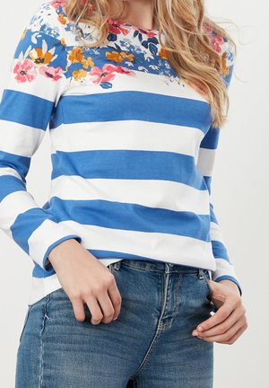 Vrouw die een shirt met lange mouwen draagt, met blauw-witte horizontale strepen en een bloemenpatroon over de schouders, gecombineerd met een blauwe spijkerbroek.