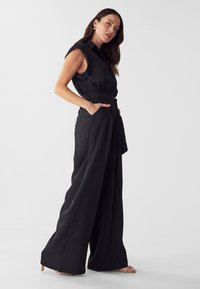 Zwarte mouwloze jumpsuit met een ceintuur in de taille, wijde broekspijpen en zijzakken. Gemaakt van zachte stof met minimale ontwerpdetaillering.