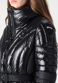 Giacca puffer nera con finitura lucida, colletto alto, design trapuntato e vita sfiancata. Presenta una patch con logo sulla spalla.