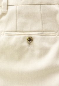 Pantalon en tissu beige avec une poche arrière à fermeture par un seul bouton et des détails de couture visibles.